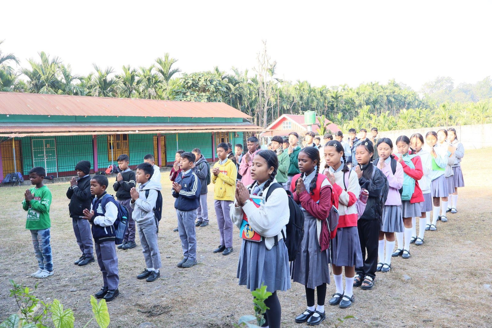 School students standing in rows in a schoolyard in front of single-story school buildings